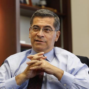California Attorney General Xavier Becerra sits with his hands and fingers crossing, explaining something.