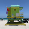 A green lifeguard hut made of wood sits on a sandy beach on a clear day. The sky is blue and people are sitting and walking on the sand,