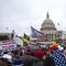 In the background is the U.S. capitol building on the day of the January 6th insurrection, with crowds visible on its steps. In the foreground are more people packed together waving Trump flags.