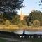 A woman is sitting on a stone wall in front of a lake as the sun sets in Essex County Branch Brook Park. There are a lot of leafy trees around and a cathedral in the far background. 