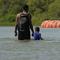 An adult and a child hold hands while they wade in water, with orange barriers floating in the background