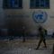 Three soldiers with guns walk in front of a beige wall with the UN emblem 