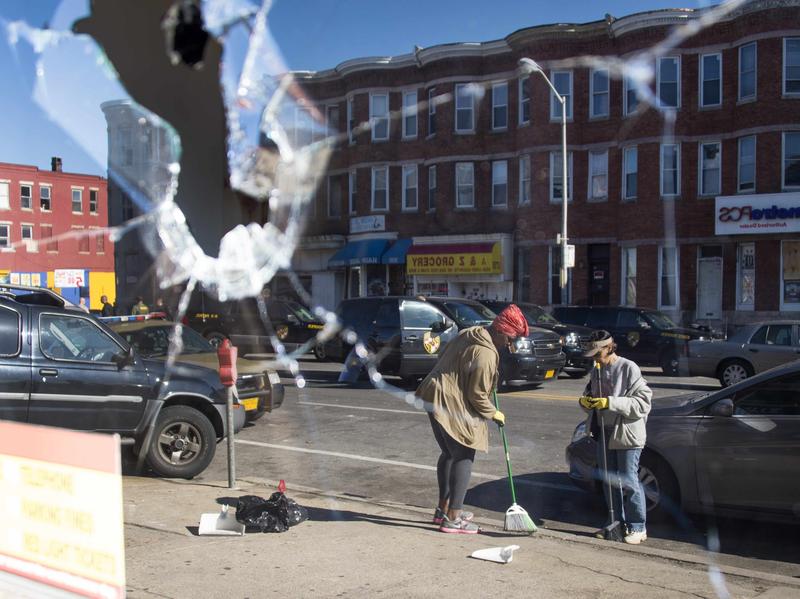 Two women sweeping up the streets are reflected in the broken window of a check cashing store in Baltimore, Maryland, on Tuesday.