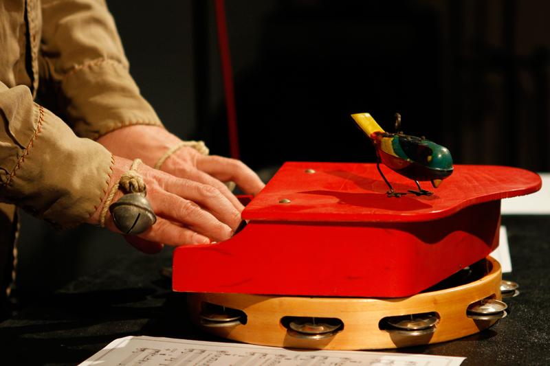 Stephin Merritt plays a toy piano, on top of a tambourine, with a mechanical toy bird.