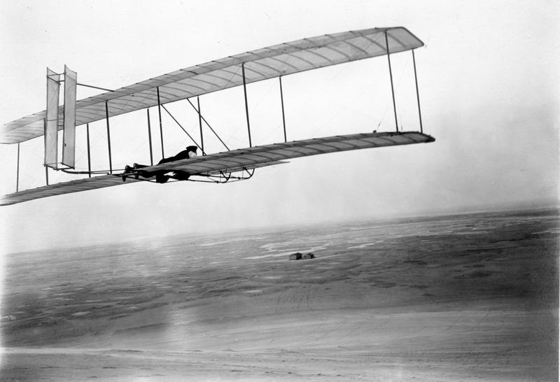 Wilbur takes wing in the 1902 glider soon after the brothers’ return to Kitty Hawk in 1903. Their camp and shed stand alone in the distant wind-swept sands.