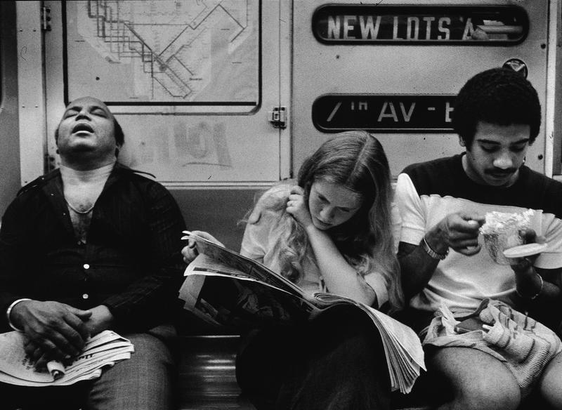 A view of three people sitting on the Number 2 line (7 Avenue Express) IRT subway, New York, 1970s. One man sleeps, a young woman reads a newspaper, and a young man eats