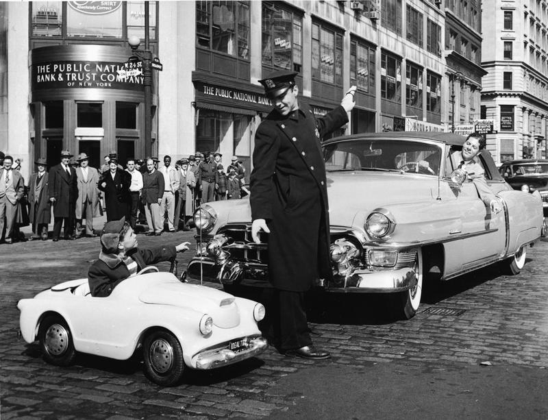 A policeman stops traffic for six year old Brian M. Dunne as the boy pilots a new fiberglass smallfry motorcar across the intersection of Broadway and 24th Street in New York, early 1950s. 