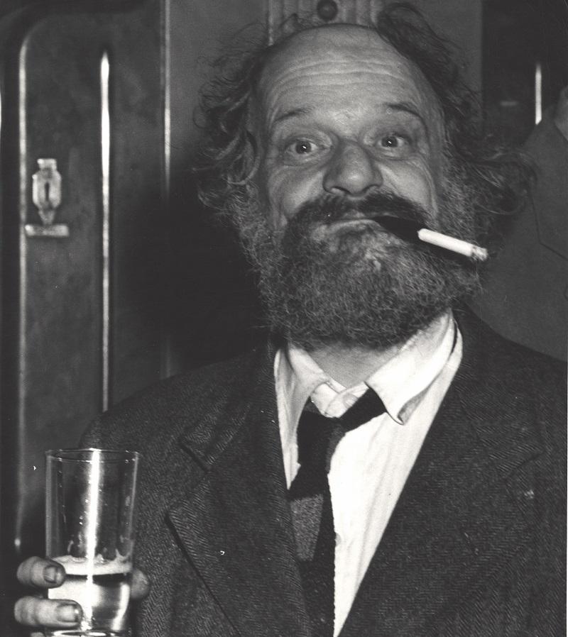 American writer and bohemian Joe Gould (1889 - 1957) holds a glass of beer and a smiles from behind a cigarette in a holder as he stands in an unidentified bar, New York, New York, 1940s or 1950s. 