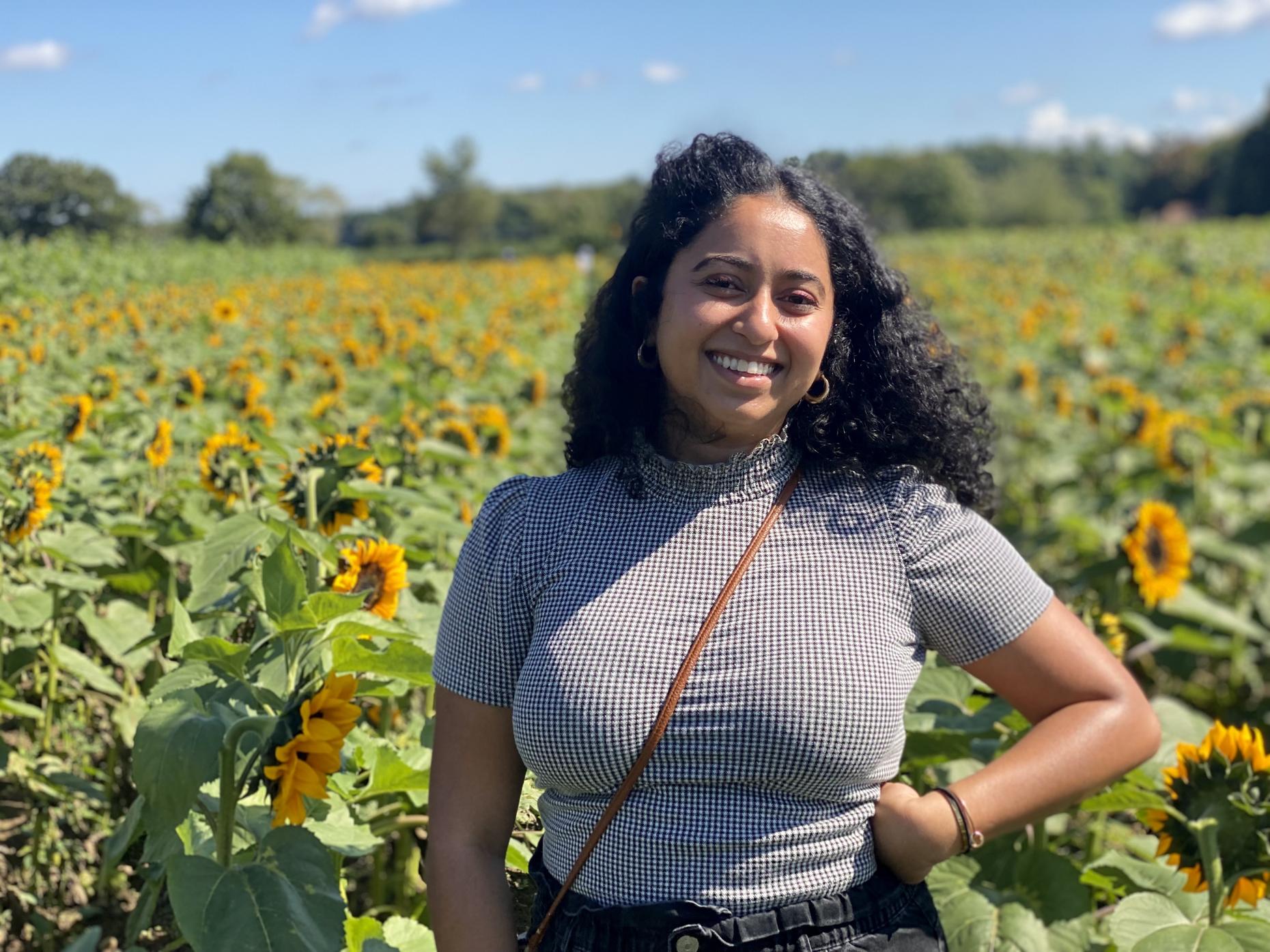 Producer Sarah Qari, smiling and standing proudly in the foreground of a field of sunflowers.